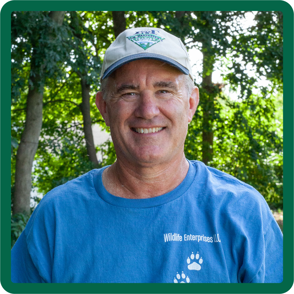Photo of Dan Lynch in a light blue Wildlife Enterprises LLC t-shirt, standing in front of sunny woods.