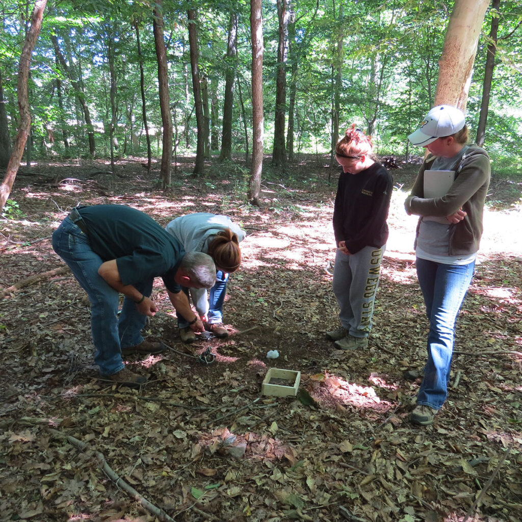 Photo of Dan and 3 students in Pennsylvania woods. Dan is showing them how to set traps.